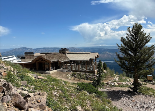 A mountain lodge with a wooden exterior, surrounded by rocky terrain and trees, overlooking a scenic valley and blue sky.